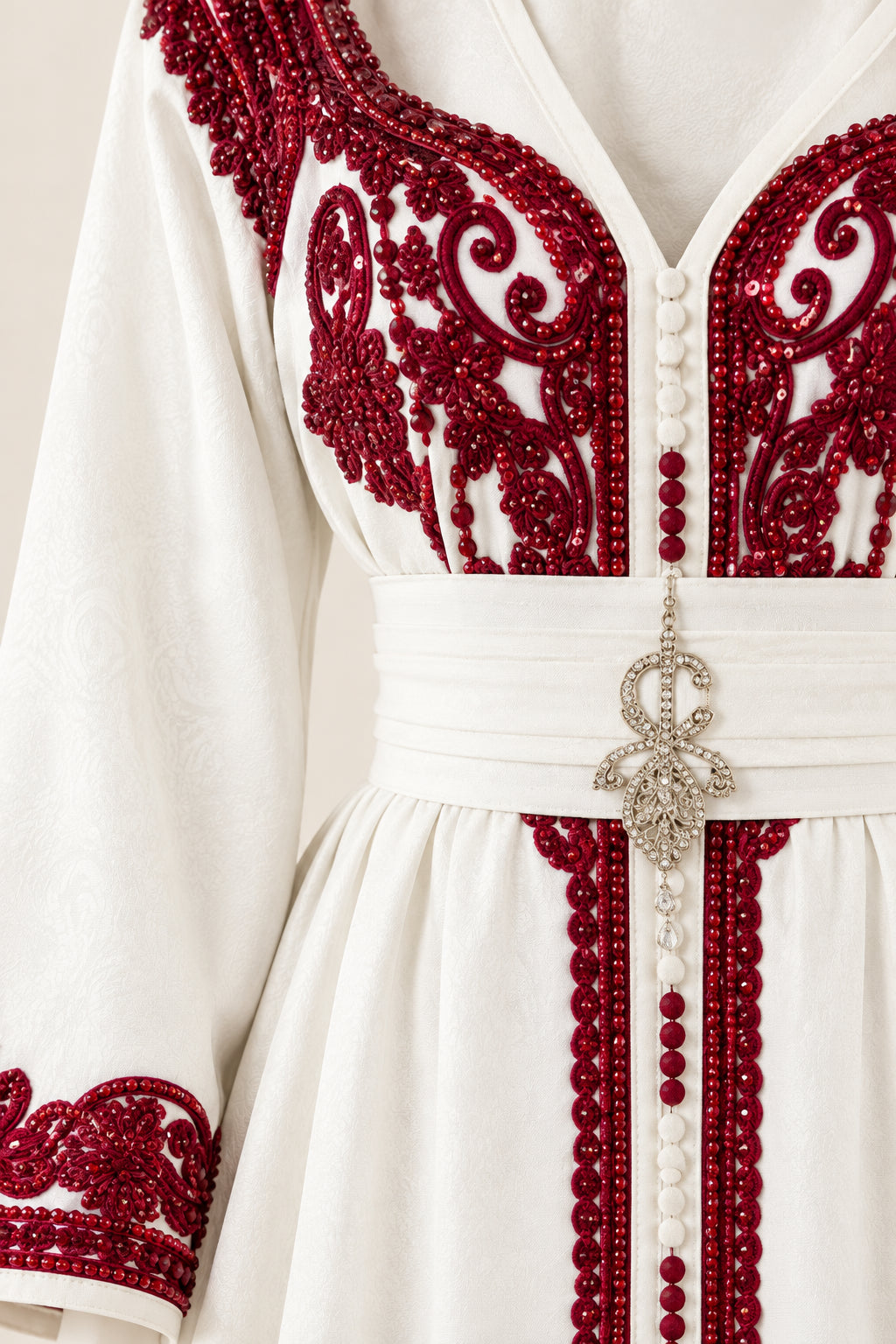 Close-up detail of ornate burgundy embroidery, beaded button trim, and waistband brooch on white kaftan