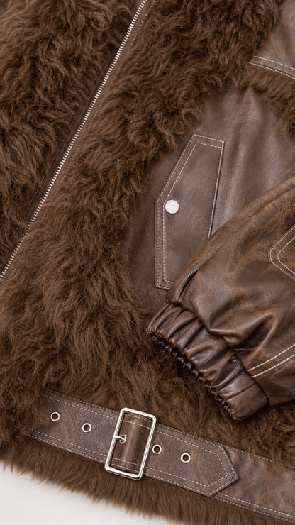 Close-up detail of brown faux leather jacket showing contrast topstitching, metal buckle belt, and textured fleece trim